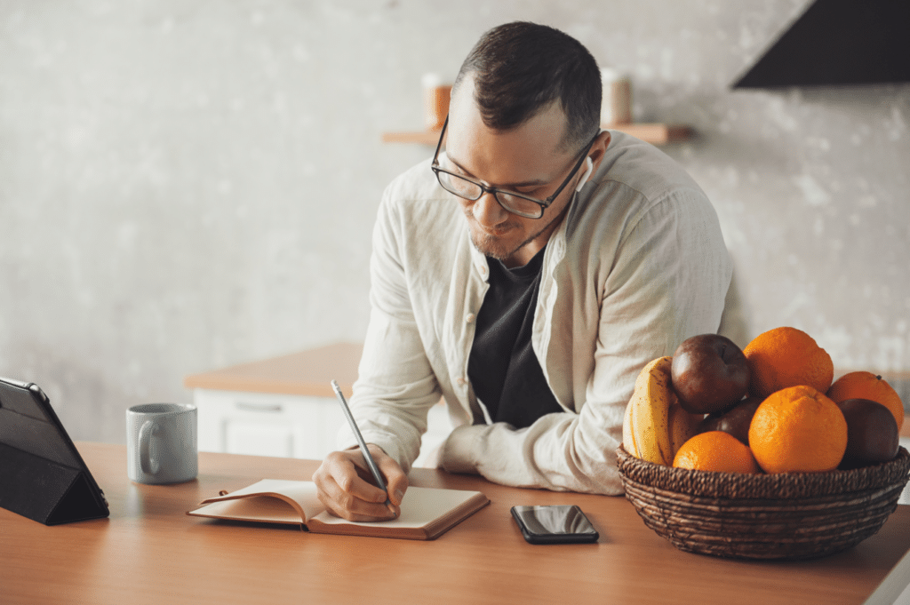 Man writing down his weight loss goals on a notebook with a bowl of fruit next to him.